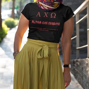 May include: A black t-shirt with a pink graphic that says "AXO Alpha Chi Omega" and the text "Life's a canvas, and I'm painting it lyre by lyre."