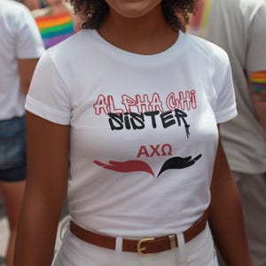 May include: White t-shirt with black and red text that says "Alpha Chi Sister AXQ". The text is in a graffiti style font. The shirt is being worn by a person with brown hair.