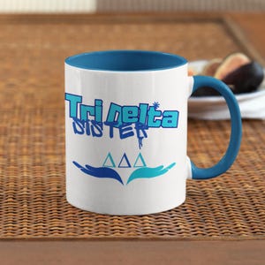 May include: White ceramic mug with a blue rim and handle. The mug features a graphic of the text "Tri Delta Sister" with three blue triangles below the text. The triangles are surrounded by two blue hands.