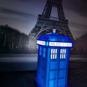 Op de afbeelding: Een blauwe Police Box-replica met de woorden "POLICE PUBLIC BOX" verlicht aan de bovenkant. Het miniatuurmodel staat tegen een achtergrond van de Eiffeltoren in Parijs. De doos heeft vier ramen en een wit licht.