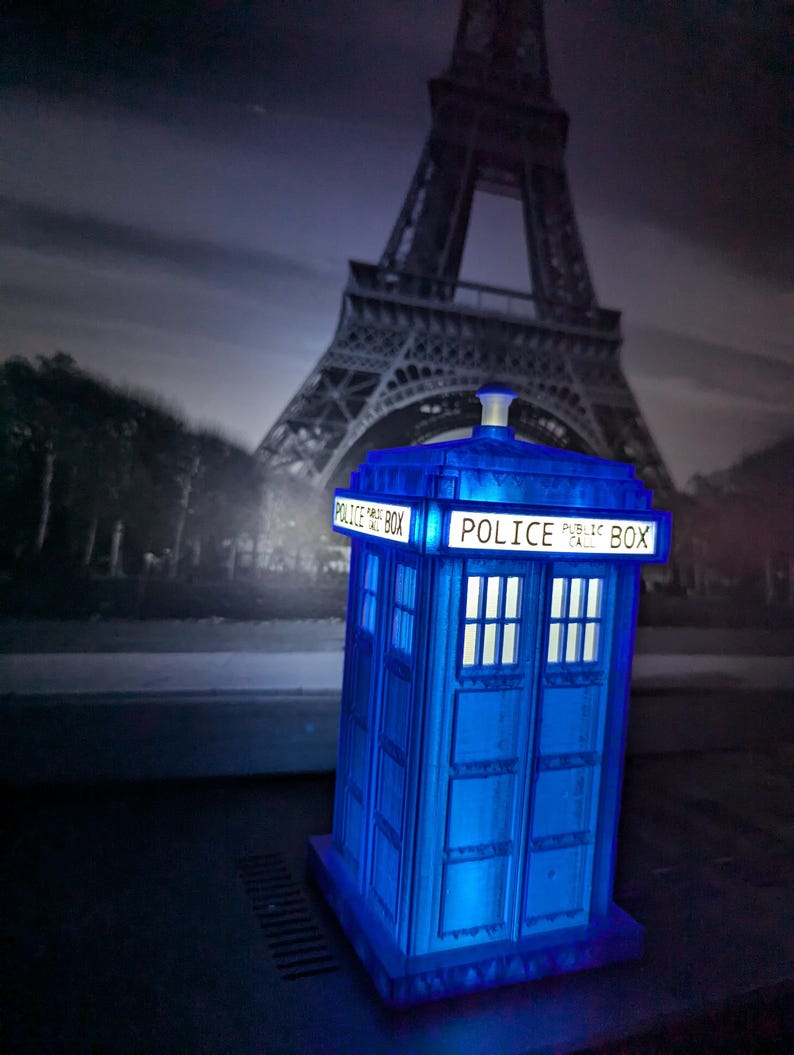Op de afbeelding: Een blauwe Police Box-replica met de woorden "POLICE PUBLIC BOX" verlicht aan de bovenkant. Het miniatuurmodel staat tegen een achtergrond van de Eiffeltoren in Parijs. De doos heeft vier ramen en een wit licht.