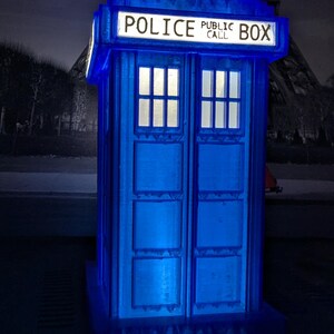 Op de afbeelding: Een blauwe, verlichte replica van een Britse politiebox, met de woorden "POLICE PUBLIC CALL BOX" bovenaan. De box heeft twee ramen en een deur en is van binnenuit verlicht. De Eiffeltoren is zichtbaar op de achtergrond.