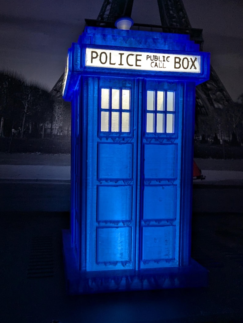 Op de afbeelding: Een blauwe, verlichte replica van een Britse politiebox, met de woorden "POLICE PUBLIC CALL BOX" bovenaan. De box heeft twee ramen en een deur en is van binnenuit verlicht. De Eiffeltoren is zichtbaar op de achtergrond.