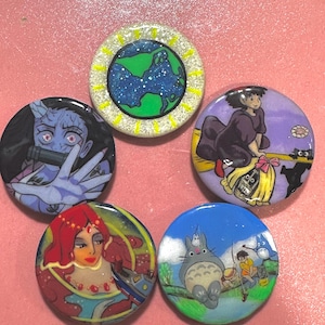 Puede incluir: Cinco botones redondos con diferentes imágenes de dibujos animados. Los botones son blancos con un acabado de purpurina plateada. Las imágenes incluyen un globo terráqueo verde y azul, una niña en una escoba, una mujer con cabello rojo, una criatura parecida a un gato y una niña y un niño pescando.