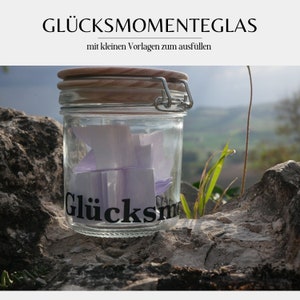 Può includere: Un barattolo di vetro trasparente con coperchio in legno e chiusura in metallo, riempito di bigliettini di carta viola piegati. Il barattolo riporta la scritta "Glücksmomente". Lo sfondo mostra un paesaggio sfocato.