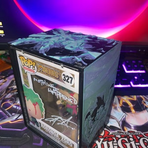 Peut inclure: Figurine Funko Pop! de Roronoa Zoro de One Piece, numéro 327, dans un étui de protection. L'étui présente un motif texturé, bleu sarcelle et noir sur le dessus et les côtés. La figurine est signée et le texte "NOTHING HAPPENED" est visible.