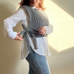 Op de afbeelding: Een grijs gebreid vest met een strik in de taille. Het vest wordt gedragen over een wit overhemd met knopen en een blauwe jeans.