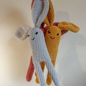 Op de afbeelding: Twee gehaakte konijnen speelgoedjes, een grijs en een oranje, met roze oren. De speelgoedjes hebben geborduurde ogen en glimlachen.