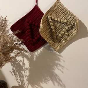 Op de afbeelding: Twee gebreide vierkanten, een rood en een beige, met een getextureerd hartontwerp. De vierkanten hangen aan een witte muur.