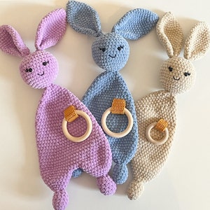Op de afbeelding: Drie gehaakte baby speelgoed konijnen met houten bijtringen. De konijnen zijn paars, blauw en beige.