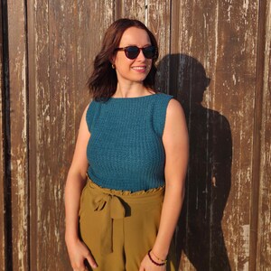 May include: A sleeveless teal blue knitted top with a boat neckline and a ribbed texture, paired with high-waisted olive green trousers featuring a fabric tie belt at the waist and dark-tinted sunglasses with a tortoiseshell frame.