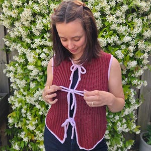 May include: A sleeveless crochet vest in a deep maroon color featuring a light lavender trim along the edges and three matching lavender tie closures down the front. The garment has a ribbed texture and a straight hemline with a V-neck opening.