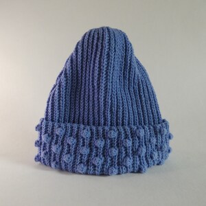 Pode incluir: Um gorro azul de malha com um design canelado. A aba apresenta um padrão texturizado de ponto de arroz. Este chapéu de inverno é um acessório aconchegante para o frio.