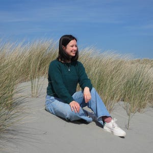 Op de afbeelding: Een persoon in een donkergroene trui en lichtblauwe jeans zit op een zandduin. De persoon draagt witte sneakers met roze sokken. Hoog, droog gras en een heldere blauwe lucht zijn op de achtergrond.