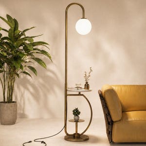 Könnte beinhalten: Eine moderne Stehlampe mit Messing-Finish, ausgestattet mit einer weißen Kugelleuchte und einem integrierten Beistelltisch mit Marmorplatte. Die Lampe hat ein kleines Regal darunter und ein schwarzes Netzkabel. Eine Topfpflanze und ein gelbes Sofa befinden sich im Hintergrund.