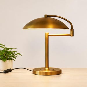 Könnte beinhalten: Messingfarbene Schreibtischlampe mit gewölbtem Schirm und verstellbarem Arm. Die Lampe hat einen runden Fuß und einen vertikalen Ständer. Im Hintergrund ist eine kleine grüne Pflanze in einem weißen Topf zu sehen. Die Lampe ist eingesteckt.