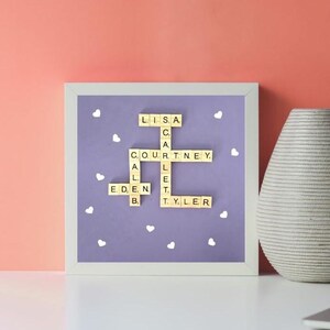 May include: A framed artwork with a lavender background features names spelled out with wooden letter tiles. White heart-shaped decorations are scattered across the background. A decorative vase and a laptop are in the background.