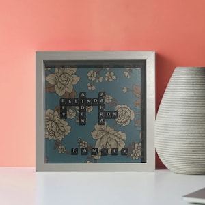 May include: A framed artwork with a blue background and a floral pattern. The artwork features a crossword puzzle made of black letter tiles that spell out the word "FAMILY" and the names "BELINDA" and "ZACHARON".