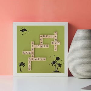 May include: A framed family name crossword puzzle with names spelled out in wooden letter tiles on a green background. The frame is white, and the background has black silhouette palm trees, birds, and a sun.