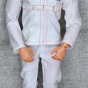May include: A vintage action figure of a man in a white jumpsuit with red stitching and a patch that says "The Six Million Dollar Man". The figure has brown hair and black shoes.
