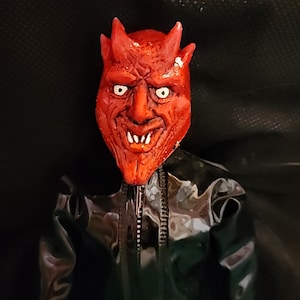 Puede incluir: Una máscara de diablo roja con cuernos y una sonrisa de dientes, usada por una muñeca vestida con una chaqueta de cuero negra.