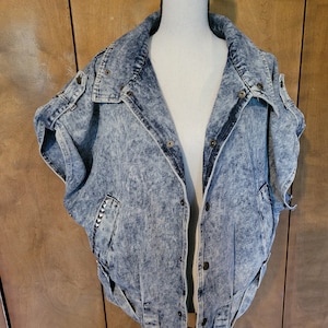 May include: A blue denim jacket with a light acid wash finish. The jacket has a stand-up collar, short sleeves, and a button closure. The jacket has a distressed look with frayed edges.