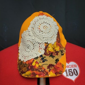 Könnte beinhalten: Orangefarbene Baseballkappe mit einem floralen Stoffpatch und zwei weißen Häkeldeckchen. Die Kappe liegt auf einer roten Oberfläche mit einem herzförmigen Etikett, auf dem "NGER 150" steht.