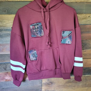 May include: Burgundy hoodie with white stripes on the sleeves and three square patches of colourful fabric sewn on the front.