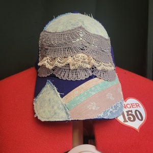 Pode incluir: Um boné de beisebol de brim azul com um design de patchwork. O boné é decorado com uma camada de renda de crochê e uma camada menor de renda branca. O boné tem um visual desgastado e é acentuado por um tecido rosa e verde com um padrão de floco de neve.