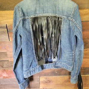 May include: A blue denim jacket with black leather fringe trim on the back and sleeves. The fringe is cut in a straight line and hangs down from the jacket.