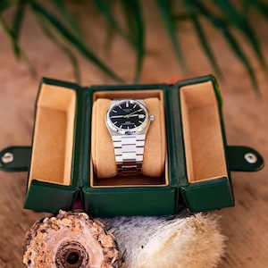 May include: A silver wristwatch with a black face and a date window is displayed in a green and brown leather watch box.