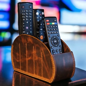 Peut inclure: Un porte-télécommandes en cuir marron avec quatre télécommandes noires à l'intérieur. Le porte-télécommandes a une forme incurvée et est fait de cuir texturé.