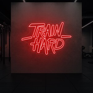 May include: A red neon sign displaying the words "TRAIN HARD" in a bold, stylized font. The sign is mounted on a textured, dark grey wall, creating a striking contrast. The image is set in a gym environment.