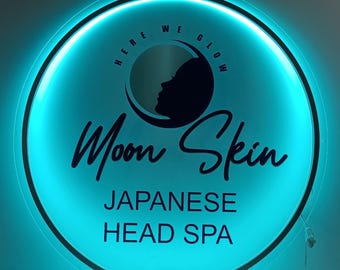 May include: A round, illuminated sign with a teal glow. The sign features the words "Moon Skin" in a cursive font, with "Japanese Head Spa" below. A silhouette of a woman's face is inside a crescent moon.