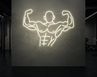 Op de afbeelding: Een neonbord met een gespierde figuur die flexeert, met de armen omhoog. Het bord is omlijnd in helder wit tegen een grijze betonnen muur, wat wijst op een sportschool- of fitnesscenterthema. De achtergrond is donker, waardoor de gloeiende figuur wordt benadrukt.