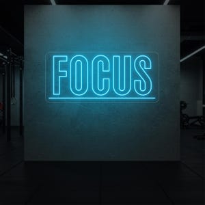 May include: A neon sign with the word "FOCUS" in bright blue against a gray wall. The sign is rectangular and the letters are outlined. The background suggests a gym or workout space.