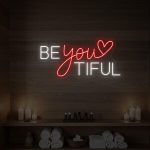 Könnte beinhalten: Ein Neonschild mit den Worten "BE youTIFUL" in Weiß und Rot. Das Wort "you" ist in einer Schreibschrift mit einem Herz anstelle des "o" geschrieben. Das Schild ist an einer Holzwand angebracht, mit Kerzen und Handtüchern auf einem Regal darunter.
