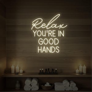 May include: A neon sign reading "Relax You're in Good Hands" in a spa environment. The sign glows with a warm white light against a wooden wall. The scene includes candles, rolled towels, and small bottles of essential oils.