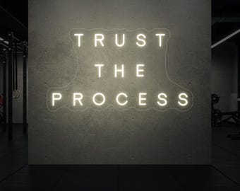 Op de afbeelding: Een wit neonbord met de tekst "TRUST THE PROCESS" op een grijze betonnen muur. Het bord bevindt zich in een sportschool, met trainingsapparatuur zichtbaar op de achtergrond.
