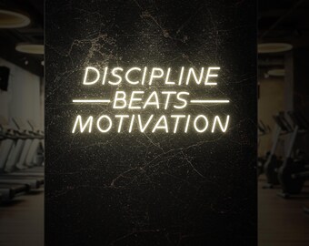 Op de afbeelding: Een neonbord met de tekst "DISCIPLINE BEATS MOTIVATION" in helderwitte letters. Het bord is bevestigd aan een donkere, getextureerde muur, mogelijk in een sportschool. Trainingsapparatuur is zichtbaar op de achtergrond.