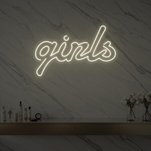 Può includere: Un'insegna al neon bianca con la parola "girls" in corsivo su un muro di marmo. Sotto, un mobile dorato ospita vasi di fiori e prodotti di bellezza. La luce brillante dell'insegna contrasta con lo sfondo.