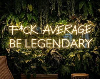 Op de afbeelding: Een neonbord met de tekst "F*CK AVERAGE BE LEGENDARY" in een cursief lettertype, verlicht in een warme witte gloed. Het bord staat tegen een achtergrond van weelderig groen gebladerte, waaronder varens en andere tropische planten. Een rieten stoel en een bijzettafel zijn zichtbaar.