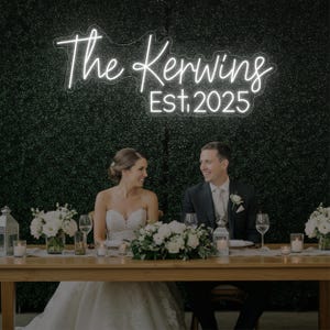 Peut inclure: Une enseigne au néon "The Kerwings Est. 2025" illumine une scène de mariage. Un couple se sourit face à face, assis à une table ornée de fleurs blanches et de bougies. L'arrière-plan est une haie verte, créant une ambiance romantique.