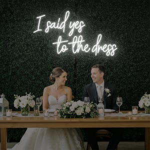 Może przedstawiać: Scena przyjęcia weselnego z panną młodą i panem młodym siedzącymi przy stole. Neonowy znak powyżej głosi "I said yes to the dress". Stół jest ozdobiony białymi kwiatami i świecami. Tło to zielony żywopłot.