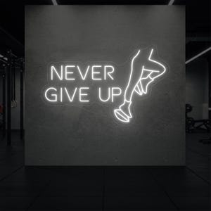 Puede incluir: Un letrero de neón blanco con las palabras "NEVER GIVE UP" y una figura corriendo. El letrero está montado en una pared de hormigón gris. El fondo es un gimnasio con equipos de ejercicio.