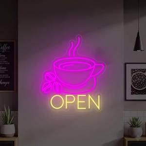 Könnte beinhalten: Ein Neonschild in Form einer Kaffeetasse mit Dampf und Kaffeebohnen, in leuchtendem Pink. Das Wort "OPEN" steht in Gelb unter der Tasse. Das Schild ist an einer grauen Wand angebracht.