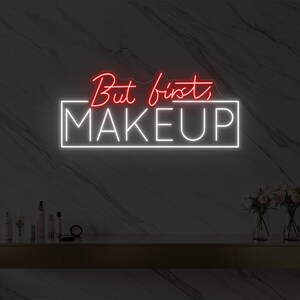 Puede incluir: Un letrero de neón con las palabras "But first, MAKEUP" en rojo y blanco. El letrero está montado en una pared de mármol sobre un tocador dorado con productos de maquillaje y flores.