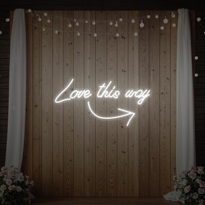 May include: A white neon sign with the words "Love this way" and an arrow, mounted on a wooden wall. The sign is illuminated, and the background includes string lights, white curtains, and floral arrangements, creating a romantic ambiance.