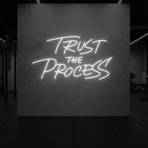 Peut inclure: Enseigne au néon blanc avec l'inscription "TRUST THE PROCESS" en lettres cursives. L'enseigne est fixée sur un mur en béton gris. L'arrière-plan est une salle de sport avec des équipements.