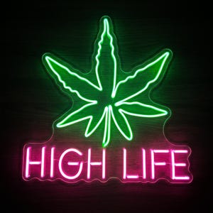 May include: A neon sign featuring a bright green cannabis leaf outline positioned above the text HIGH LIFE illuminated in vibrant pink neon tubing. The sign is mounted on a clear backing and glows against a dark, non-descript background.